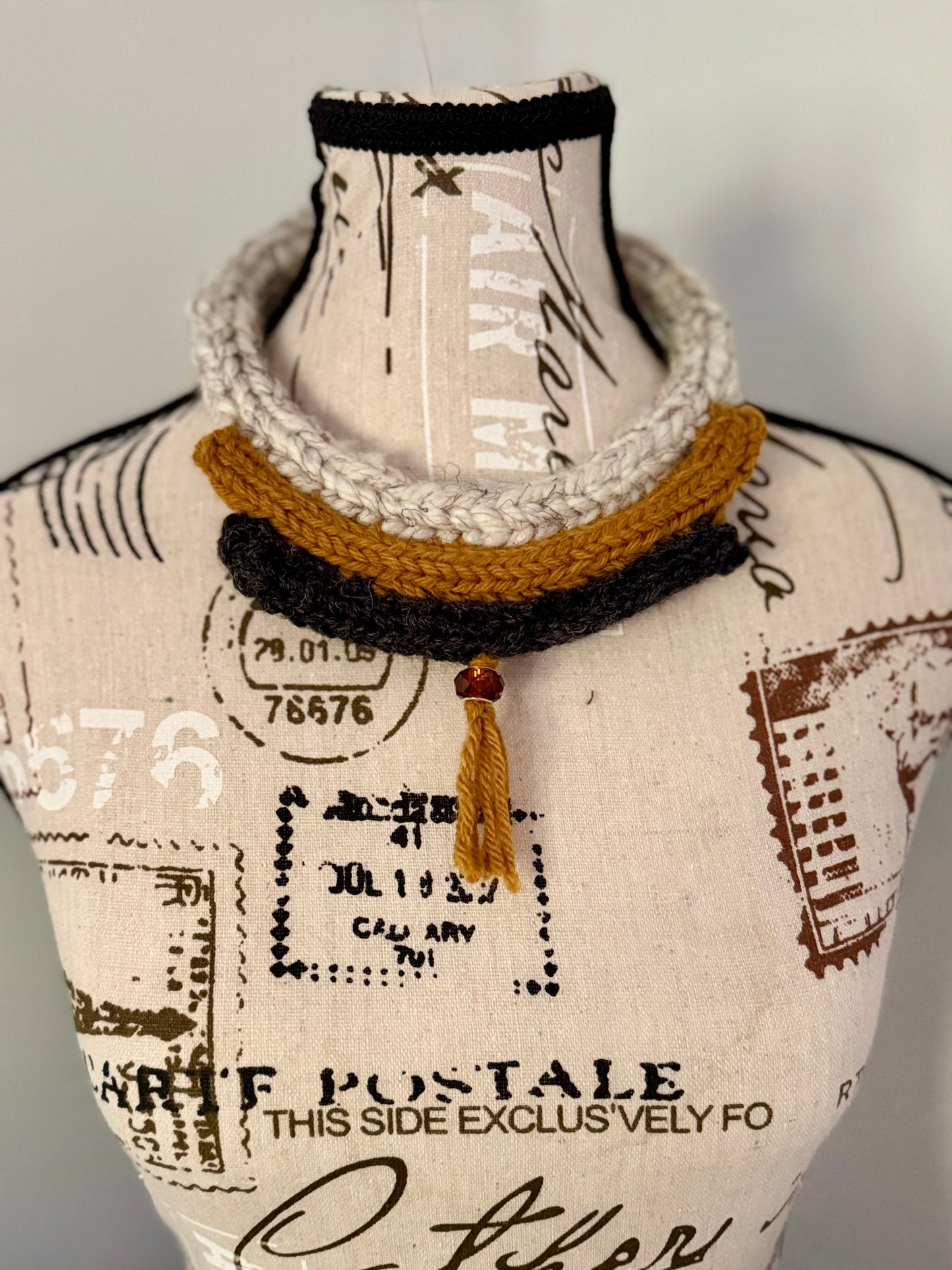 Hand knitted white, gold and black icords form a structured and elegant necklace featuring a single centered tassle with a brown bead accent.