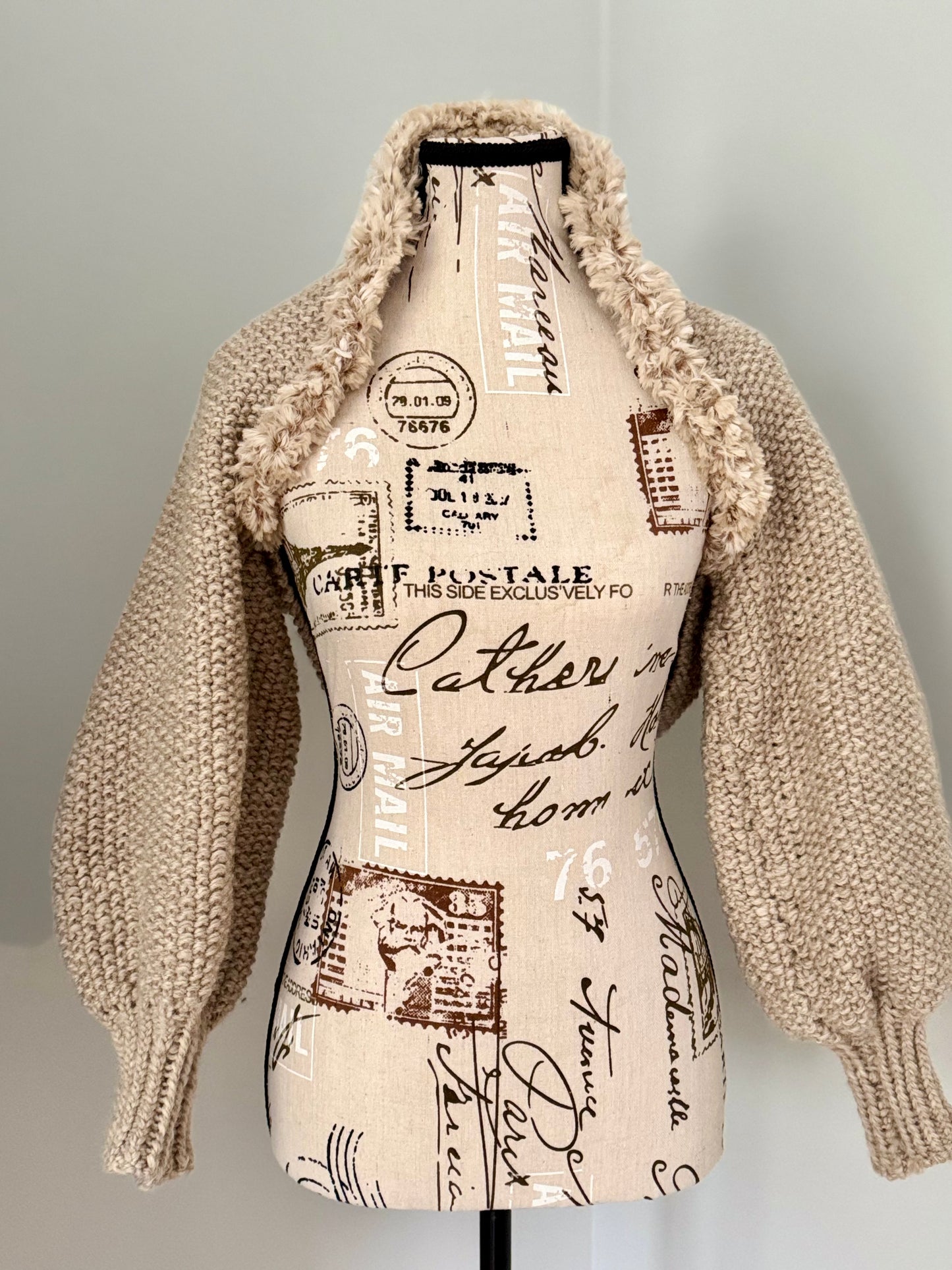 Structured hand-knitted beige shrug featuring seed stitch faux fur trim and prominent cuffs.