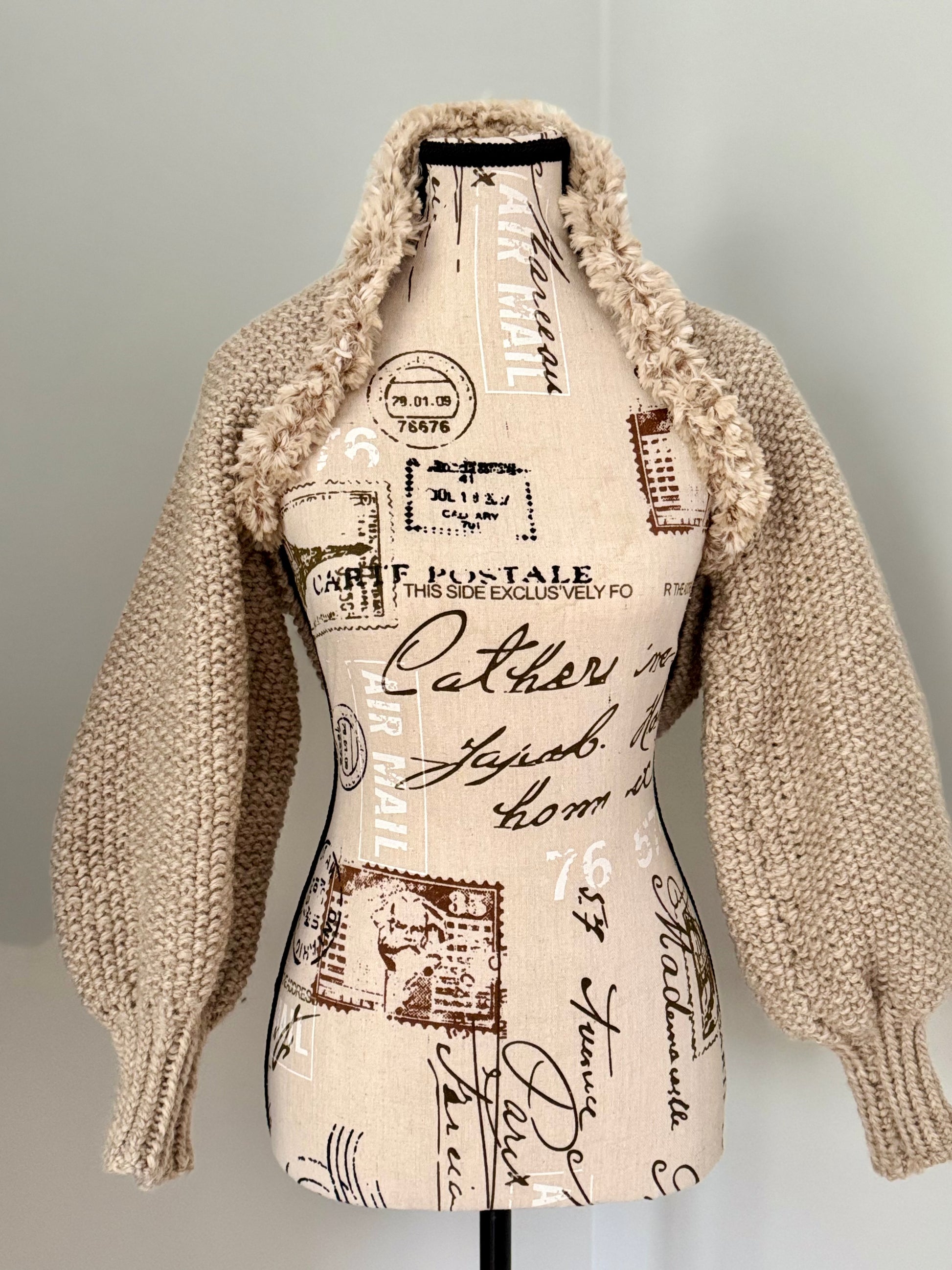 Structured hand-knitted beige shrug featuring seed stitch faux fur trim and prominent cuffs.