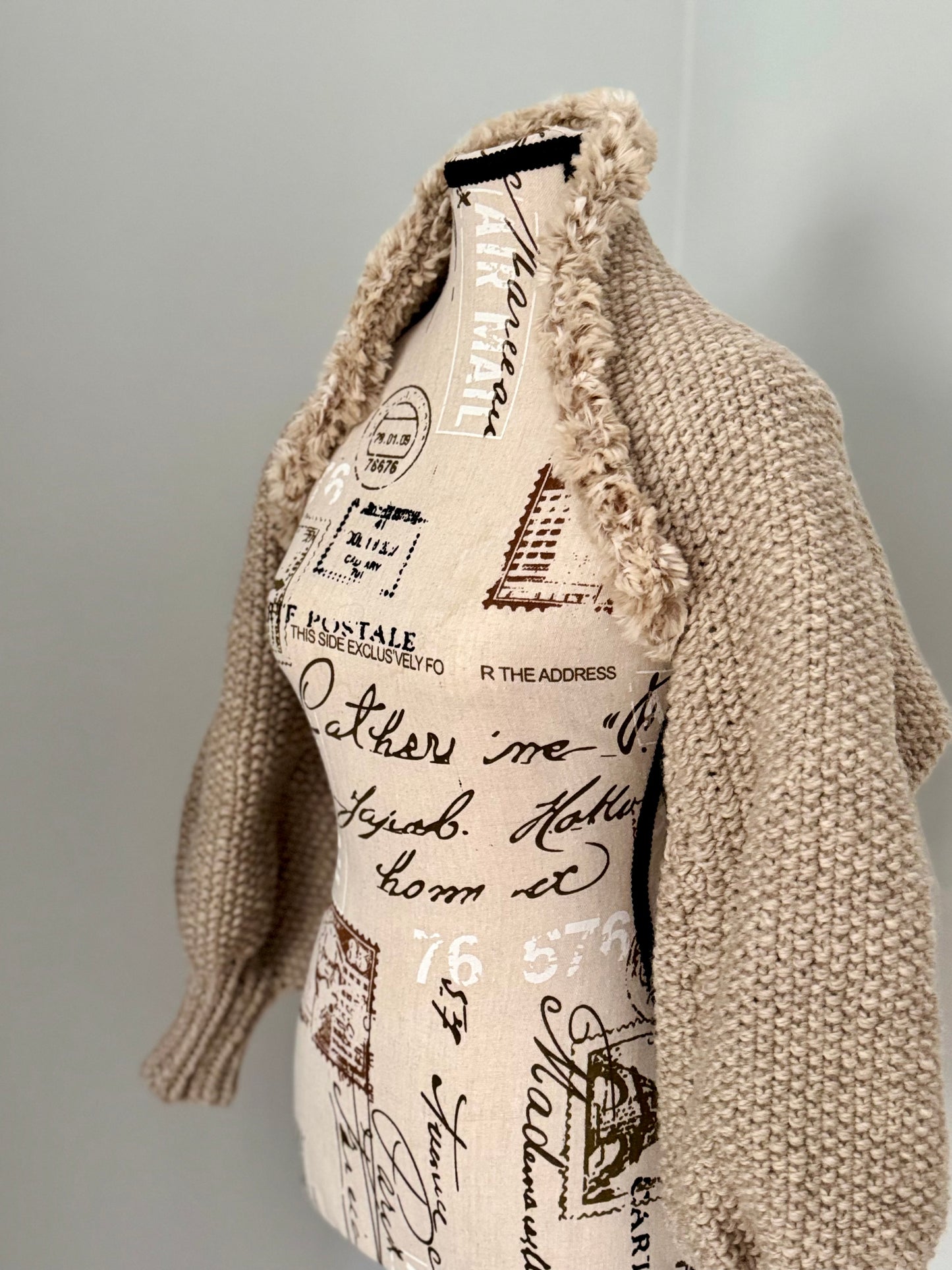 Structured hand-knitted beige shrug featuring seed stitch faux fur trim and prominent cuffs.