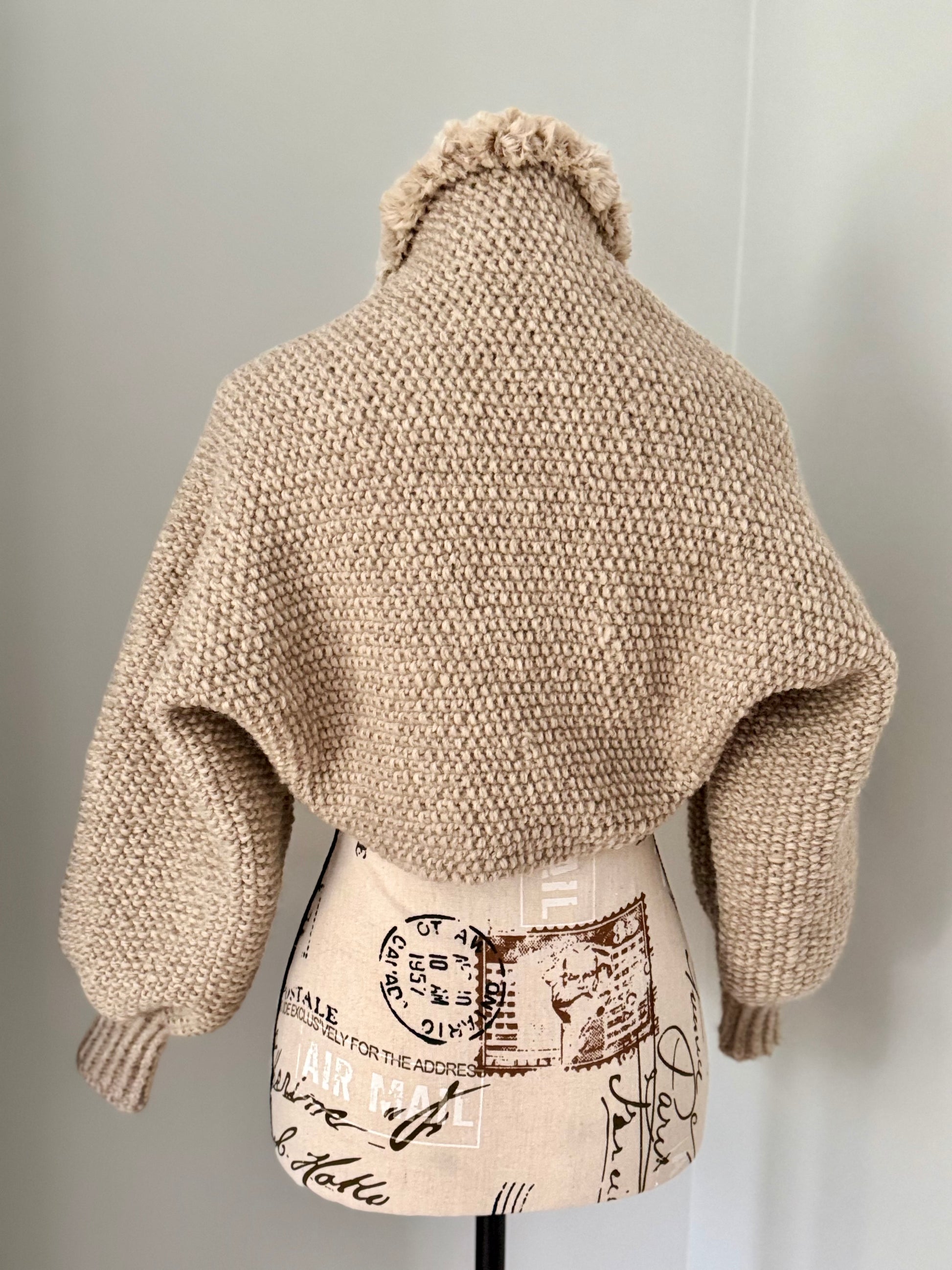 Structured hand-knitted beige shrug featuring seed stitch faux fur trim and prominent cuffs.